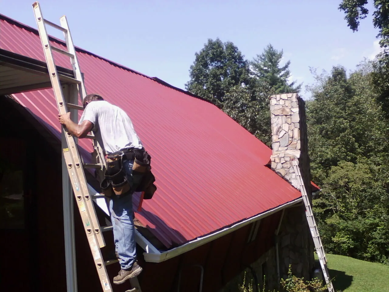 Roofer on a ladder beside a red metal roof and stone chimney — Soffit & Fascia Repair by Roofing Brothers in Homer Glen